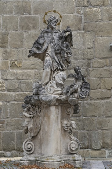 Baroque stone statue of a saint with angel on a decorated pedestal in front of a stone wall, parish