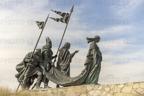 Sculpture group of historical figures with flags on a stone platform under a blue sky,