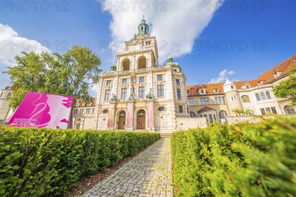 Historic building with towers and pink banners framed by manicured hedges and blue sky, Munich,