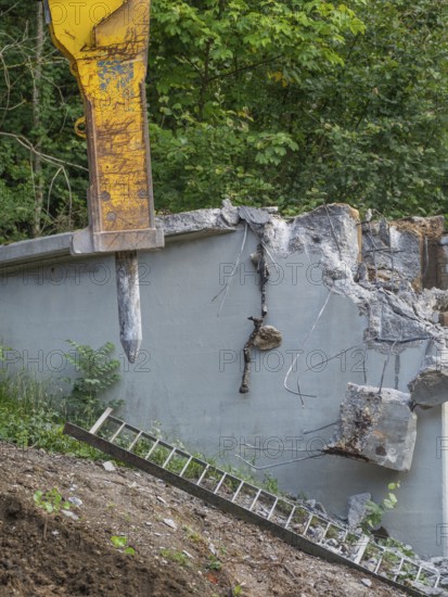 A demolition hammer destroys concrete, a ladder leans against the wall, surrounded by plants,