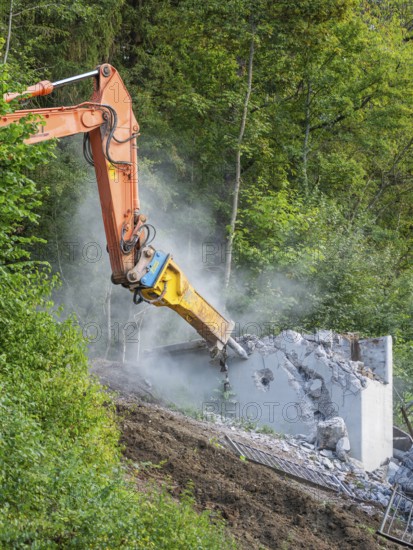An excavator breaks a concrete wall in a green environment, dust rises, bridge demolition, Calw,