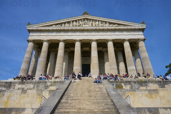 Temple-like structure with monumental columns and visitors on a staircase under a blue sky,