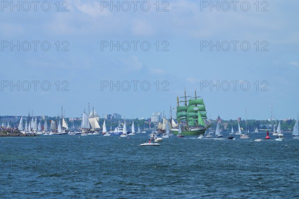Green tall ship Alexander von Humbold 2 surrounded by many boats in front of an urban backdrop,