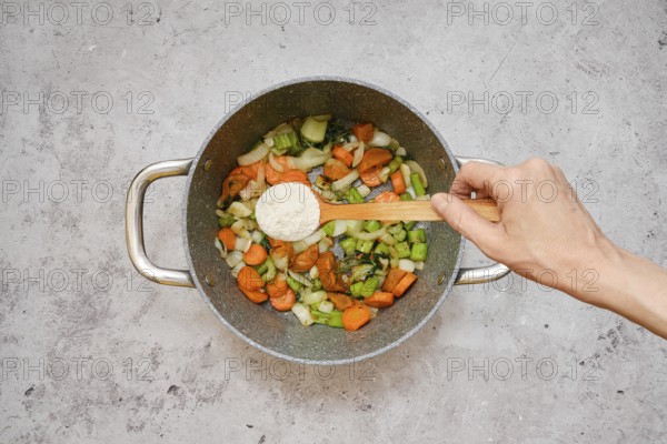 Freshly chopped carrots, celery, and onions are fried in a pot. A hand is sprinkling flour over the