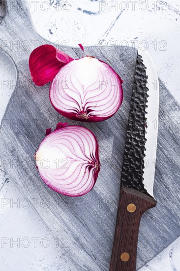 A vibrant red onion is cut in half, on a rustic wooden chopping board beside a sharp knife, ready