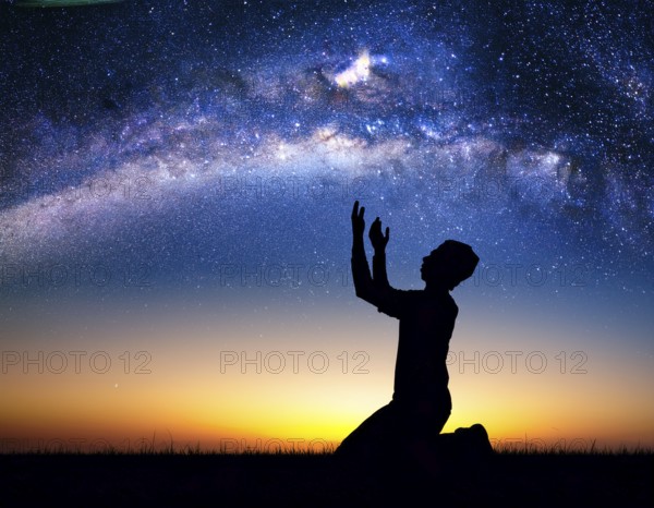A man kneels in peaceful reflection under the vibrant Milky Way galaxy at sunset in an open natural