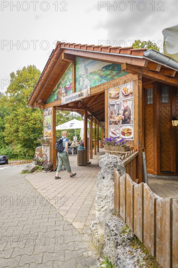 A wooden restaurant at the edge of the forest with passing hikers on a paved path, Lichtenstein