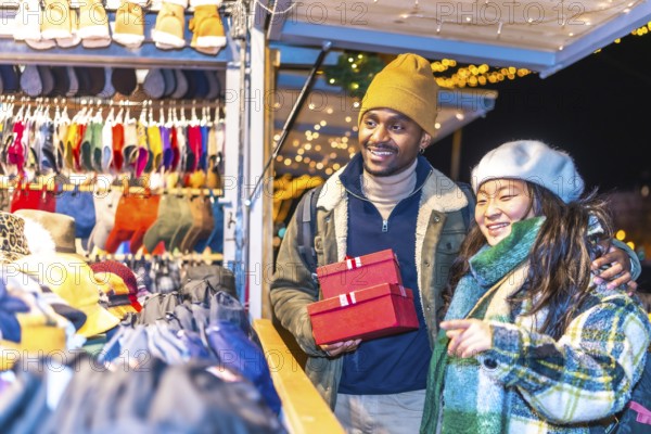 Young multiethnic couple enjoying christmas shopping at a decorated christmas market, holding red