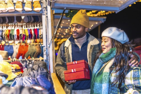 Young multiethnic couple enjoying christmas shopping at a decorated christmas market, holding red