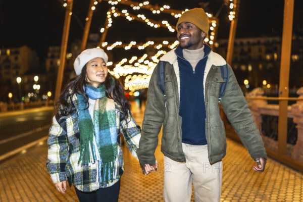 Young multiethnic couple holding hands and walking under christmas lights decorations, enjoying