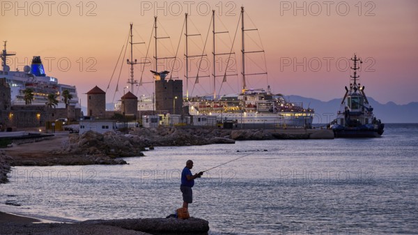 A fisherman fishing at sunset at an illuminated harbour with several ships on the horizon, Rhodes