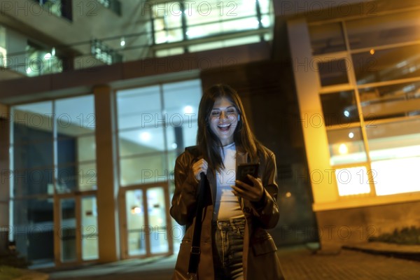 Young woman standing on an urban street at night, happily looking at her smartphone while holding a