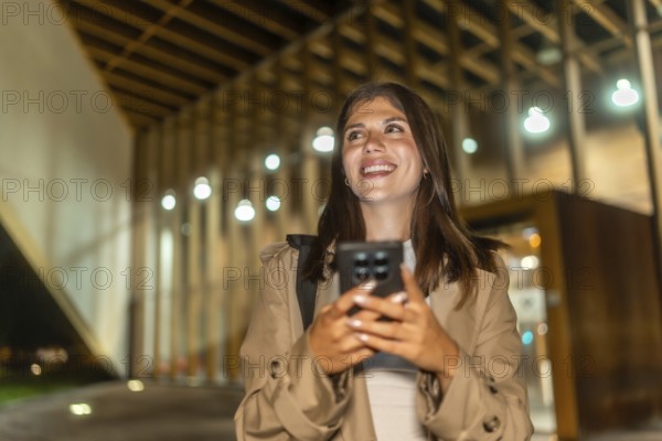Young woman smiling, holding phone while walking illuminated city streets at night, confidently