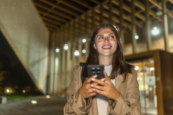 Young woman smiling while holding a smartphone and looking up, exploring a modern city street at