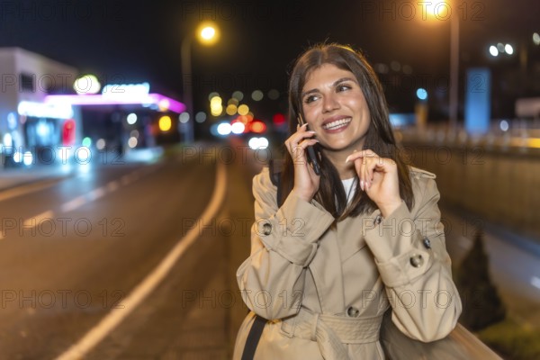 Young woman in a trench coat smiling as she talks on her mobile phone on a bustling city street at