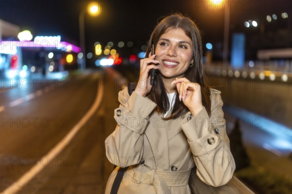 Young woman in a trench coat smiling happily while having a phone call, standing on a city street