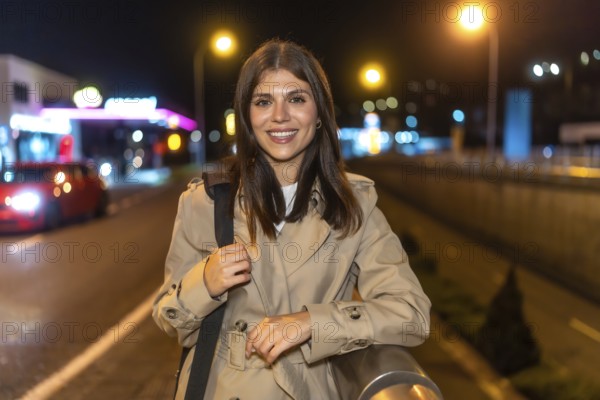 Young woman smiling while standing in a trench coat on an urban street at night, illuminated by