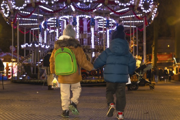 Two young brothers, holding hands, walk away from a brightly lit carousel at a christmas market in