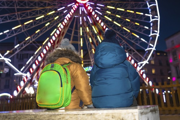 Two children with winter coats and backpacks admiring a brightly lit ferris wheel at a christmas