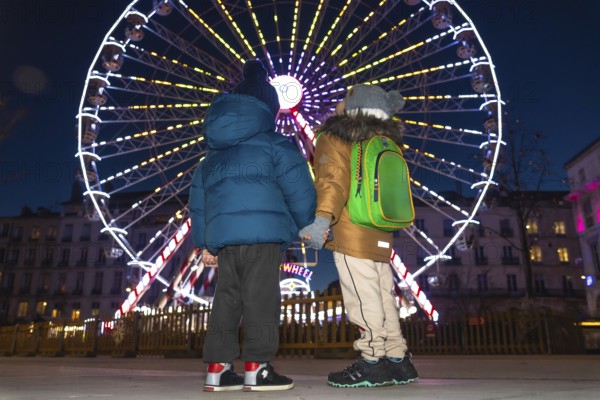 Two children holding hands are admiring a brightly lit ferris wheel at a christmas market in