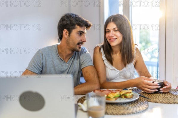 Young couple talking and having breakfast in their kitchen, with a laptop on the table and natural