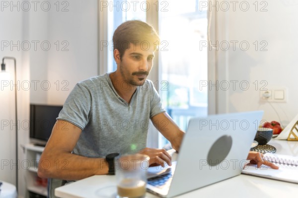 Young man working from home, typing on a laptop and jotting down notes while basking in the warm