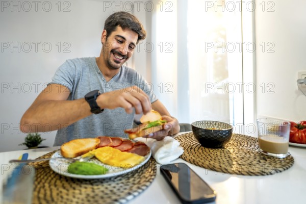 Young man smiling while preparing a delicious sandwich for breakfast at home, enjoying a healthy