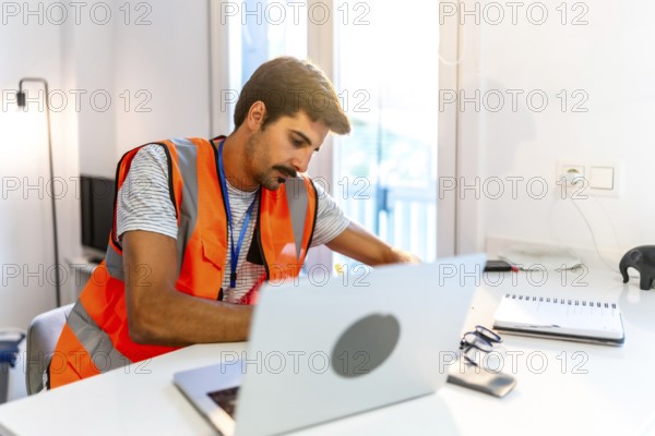 Focused engineer wearing a high visibility vest working from home, using a laptop and taking notes