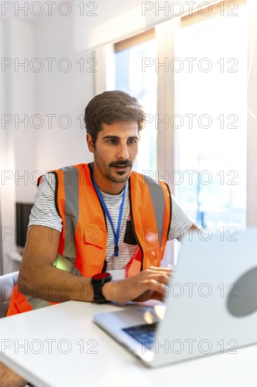 Focused engineer wearing high visibility vest working on laptop in bright modern office, managing