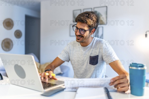 Young man working comfortably at home, engaging with a laptop and reviewing documents, smiling