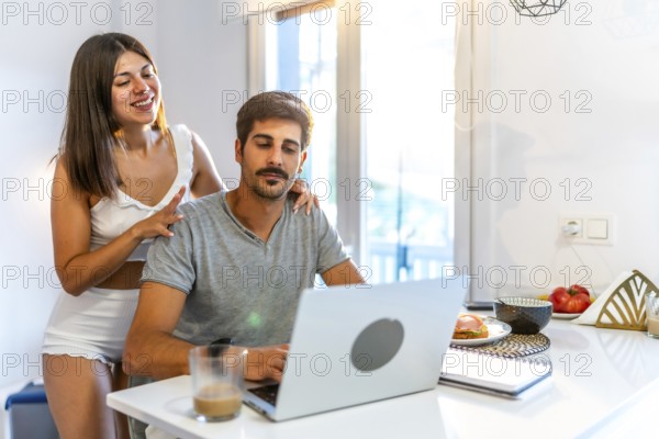 Young woman massaging her husband's shoulders while he's working from home on his laptop in the