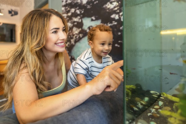 Happy mother and her baby son pointing at colorful fish swimming in a home aquarium, enjoying