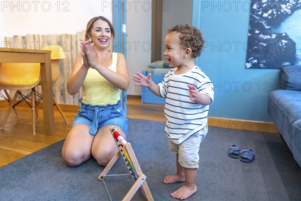 Mother encouraging her baby boy to walk, clapping hands in living room, promoting child development