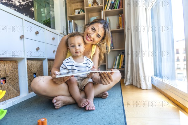 Happy mother and her baby son reading a book together, sitting on the floor in the living room at