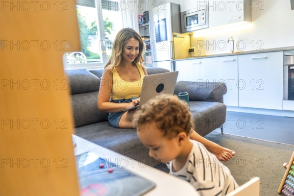 Woman telecommuting on her laptop while her baby plays nearby in a modern living room, blending