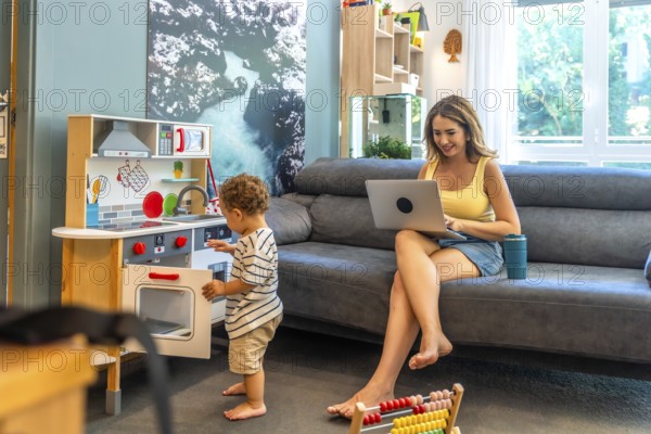 Woman working remotely on laptop while her toddler plays with a toy kitchen, demonstrating work