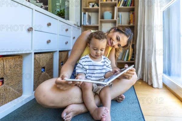 Young mother sitting on the floor reading a book with her little son, enjoying quality time