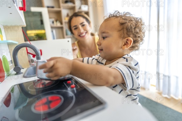 Happy toddler playing with toy kitchen while his mother watches him, promoting early childhood