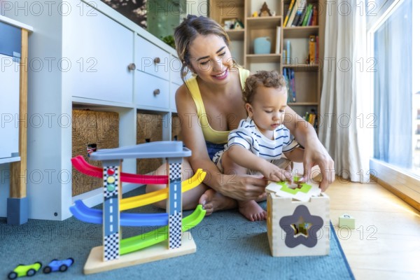 Happy mother helping her baby son playing with a shape sorter toy, sitting on a play mat in a