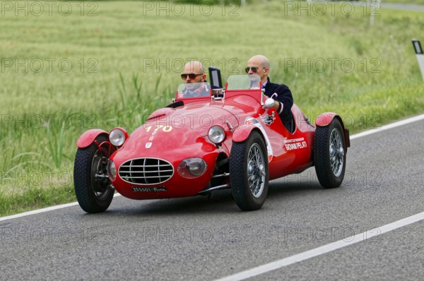 An open red sports car with two drivers drives through a green landscape, classic car, car race,