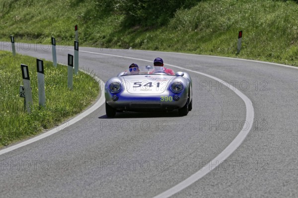 A silver racing car with two people drives swiftly along a winding road, classic car, car race,