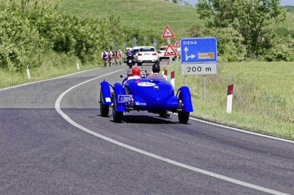 Blue classic car drives towards Siena, accompanied by other vehicles and signs, classic car, car