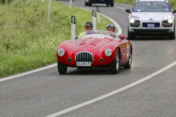 A red vintage racing car drives along green fields on a winding country road during a rally,