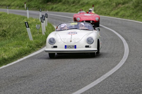 A white Porsche convertible in classic car style drives on a winding country road in front of a red