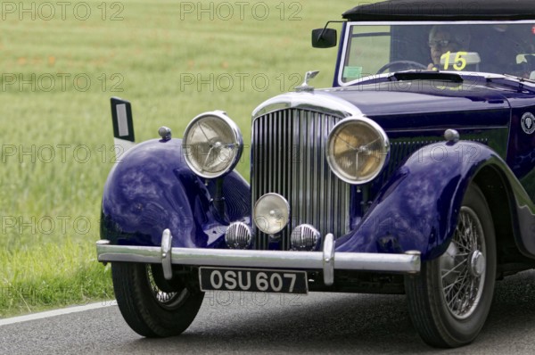 An elegant purple vintage car on a road, highlighted front headlights, surrounded by green fields,