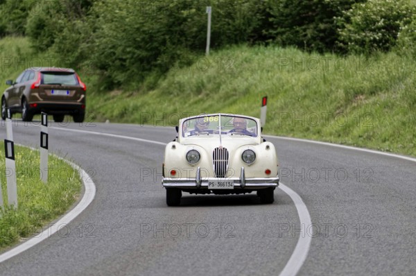 A classic convertible drives along a winding country road surrounded by green countryside, classic
