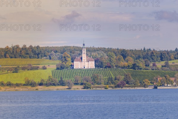 A church on a hill surrounded by vineyards and forest, with a view of a lake under a colorful