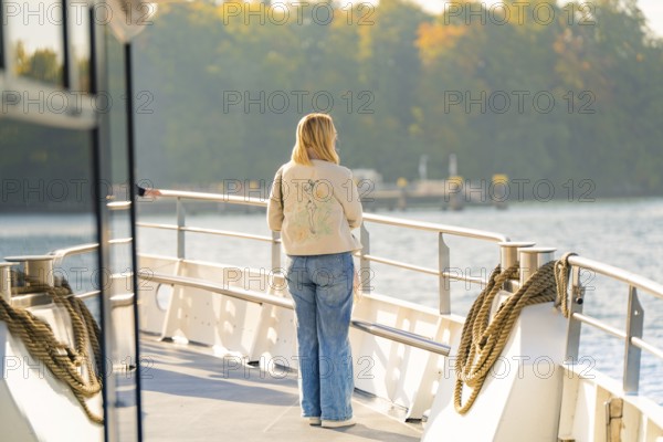 Woman on the deck of a boat looking at water and autumn trees in the distance, boat trip, Lake