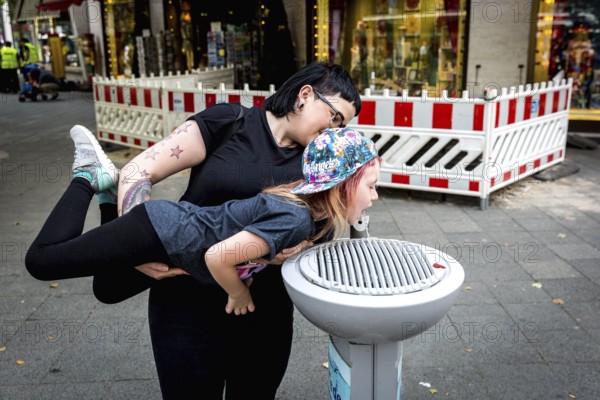 DEU Germany Germany Berlin A woman holds a girl over a stream of water gushing from a public water