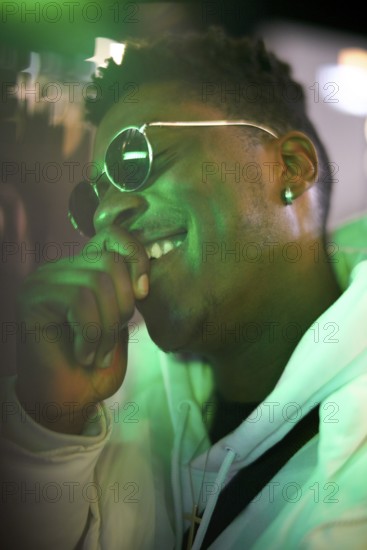 Greenlit African man wearing sunglasses at night as shade of anonymity, laughing, happy, giggle,
